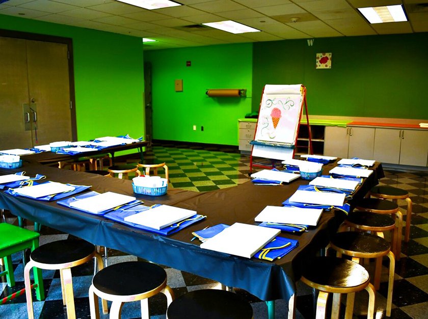 A green room with tables set up in a U shape facing a large aisle, and at each seat of the tables there is a canvas and apron.