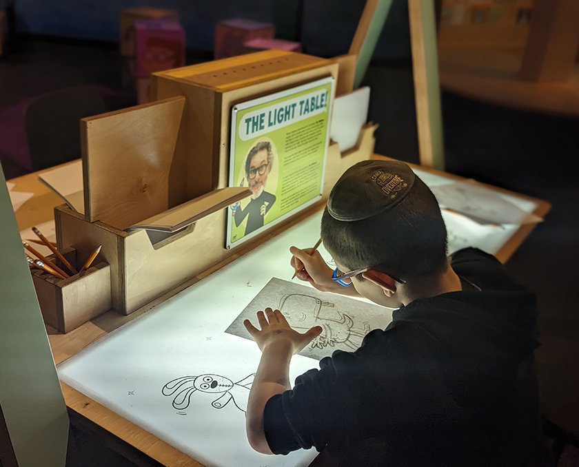 A child sitting at a light box table in the Mo Willems exhibit tracing images of Mo Willems characters. 