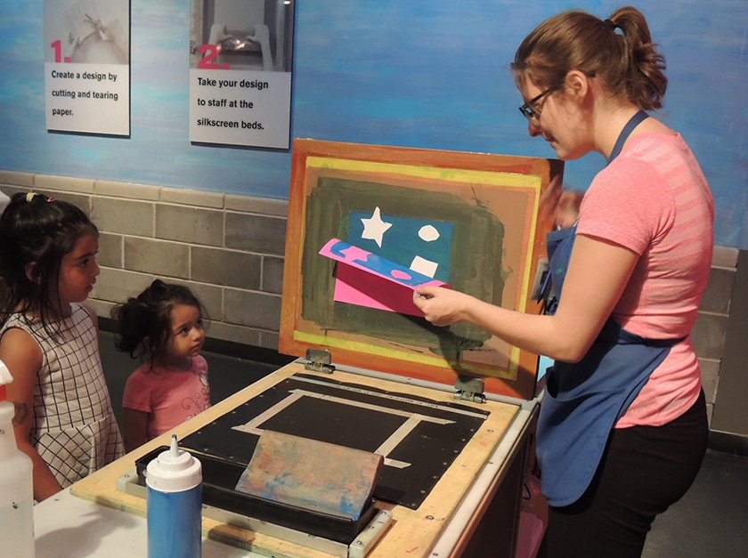 An educator showing two young visitors how to create screen-printed work. 