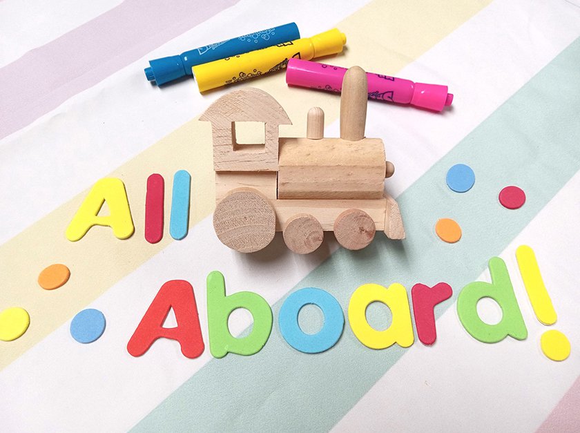 A wooden toy train, with a blue, yellow and pink marker above it, and colorful text "All Aboard" to the left and under the train. 