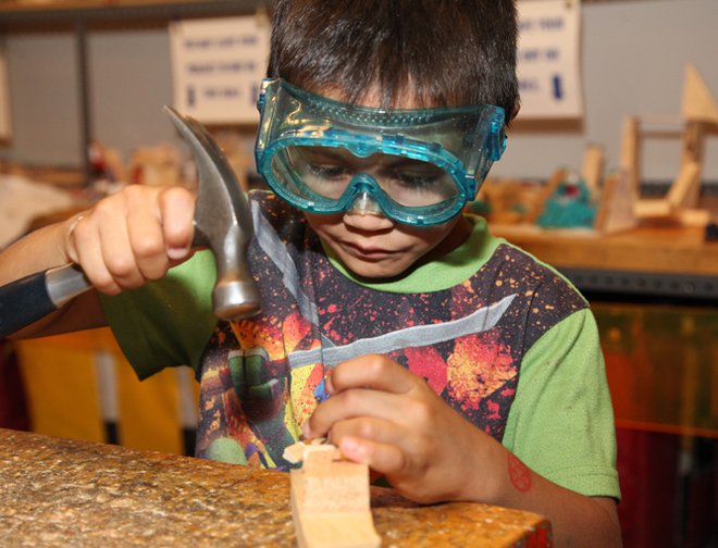 Boy using a hammer and nail to create wooden project while wearing safety googles. 