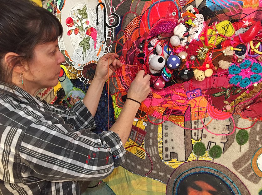 Artist Julie Peppito standing in front of unfinished canvas featuring discarded toys and mixed media textiles.