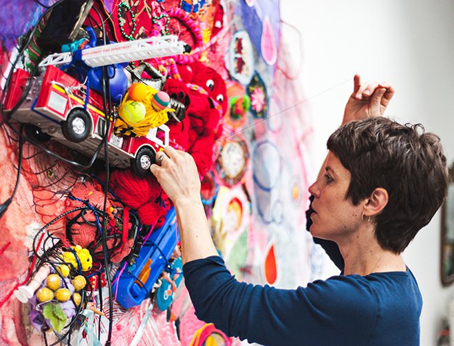 Artist with sewing needle adding brightly colored fabric and discarded toys to a canvas.