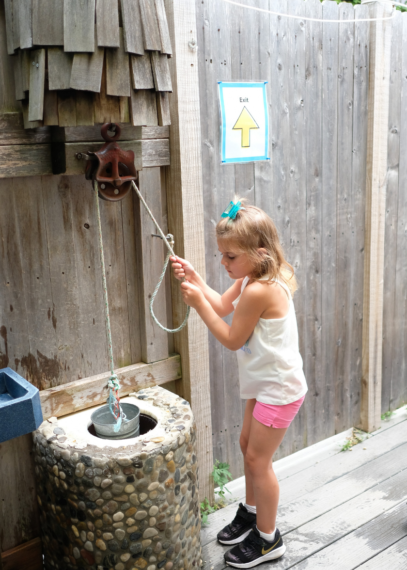 A child standing in the H2Oh! area using a pulley system to get water in a metal bucket from a small rock well.