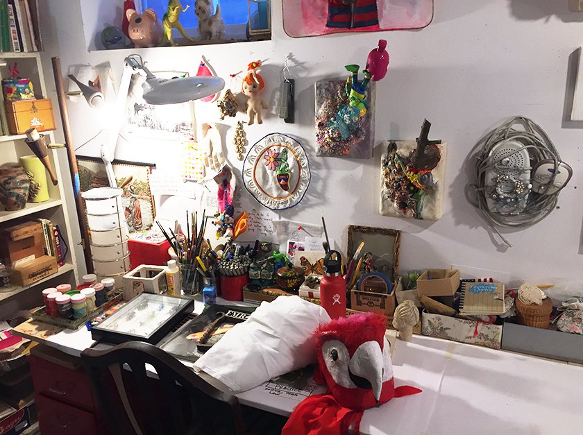 Drawing table filled with brushes, markers, paint pots and surrounded by discarded toys and miniature textile panels