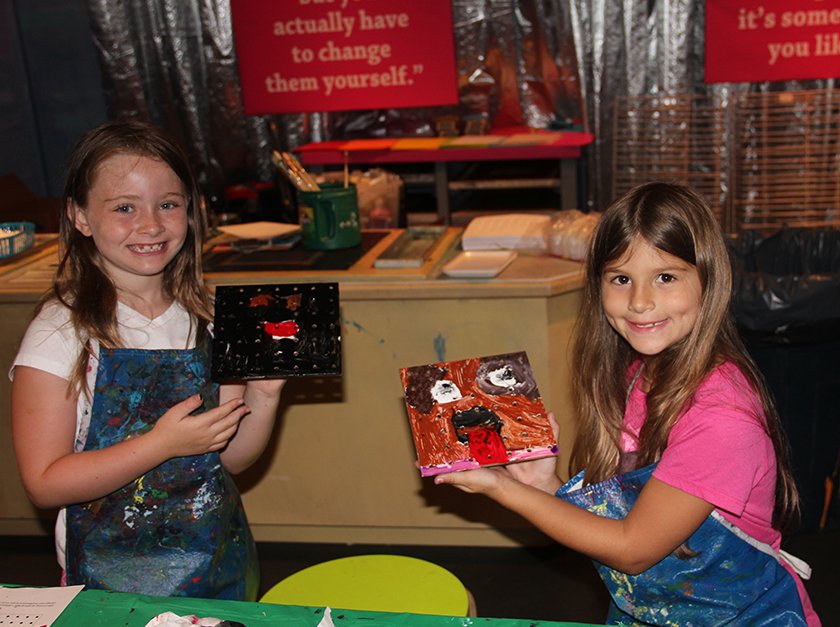 Two girls in paint-filled smocks showing off their paintings. 