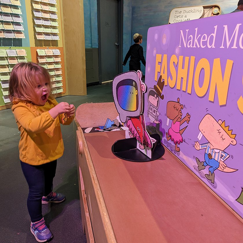 A child  with a happy expression in the Mo Willems exhibit holding small pieces of felt clothing to place on a felt naked mole-rat. 
