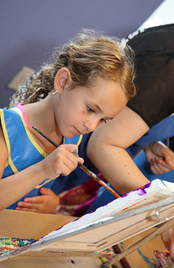 Child wearing an apron while painting a canvas on an easel. 