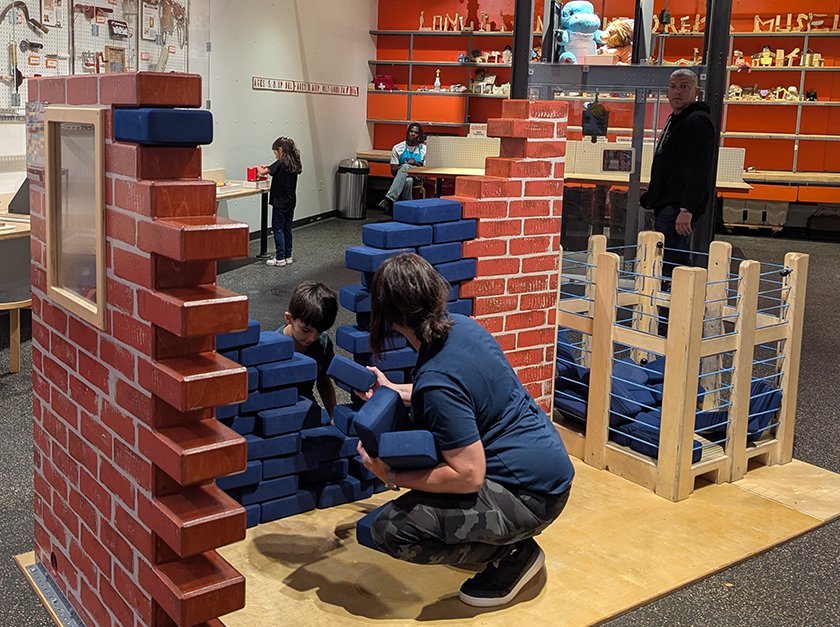 A child and adult building a large brick wall with blue yoga blocks in the front of the Tool Box exhibit. 