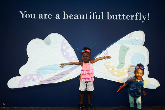 A child standing in front of large butterfly wings with her hands outstreched and another child standing to the right of her with text "You are a beautiful butterfly" above. 
