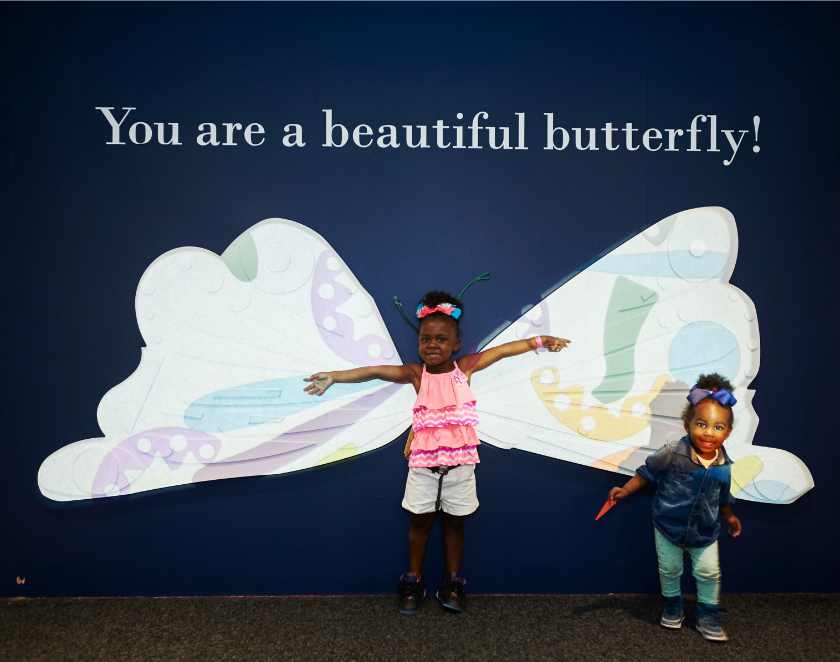 A child standing in front of large butterfly wings with her hands outstreched and another child standing to the right of her with text "You are a beautiful butterfly" above. 
