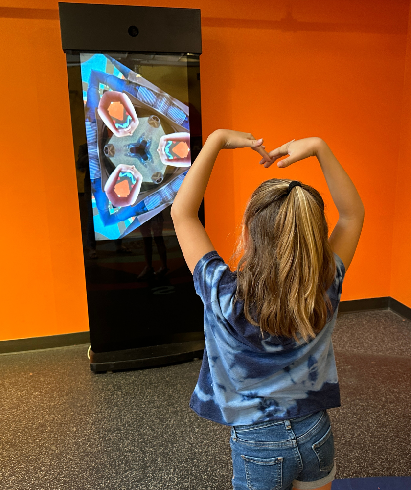 A child standing in front of a kalediscope screen with her arms above her head in a heart shape. 