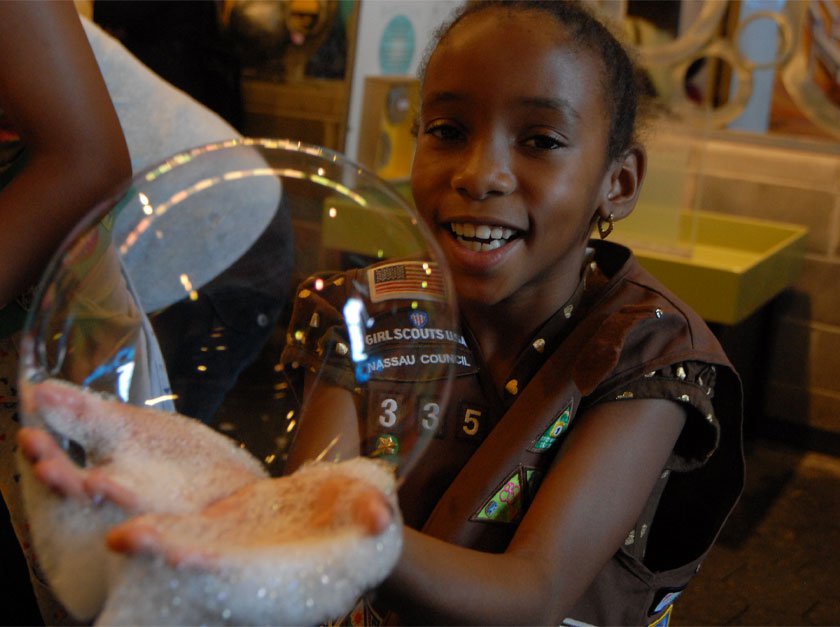 A child wearing a Brownie Vest in the LICM Bubbles exhibit with suds on their hands holding a bubble. 