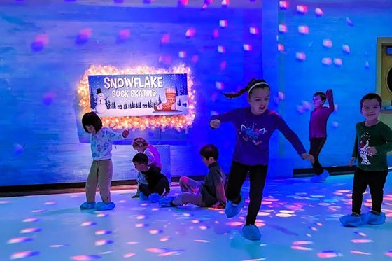 A group of kids wearing covers over their shoes while 'skating' on synthetic ice with signage behind them "Snowflake Sock Skating." 