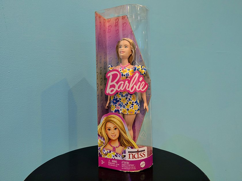 A Barbie with Down Syndrome wearing a short dress with blue and yellow flowers still in the original packaging. 