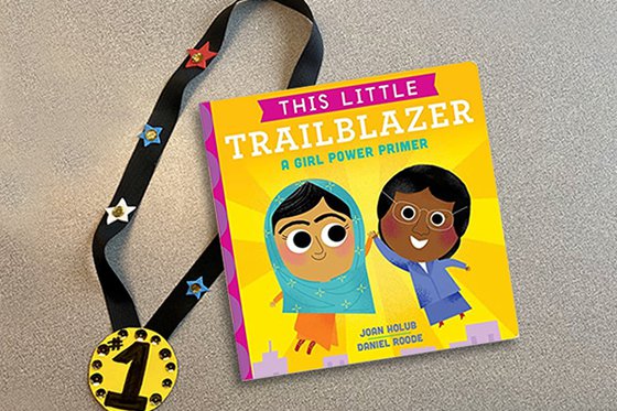 The book "This Little Trailblazer: A Girl Power Primer" and a handcrafted #1 gold medal lanyard. 