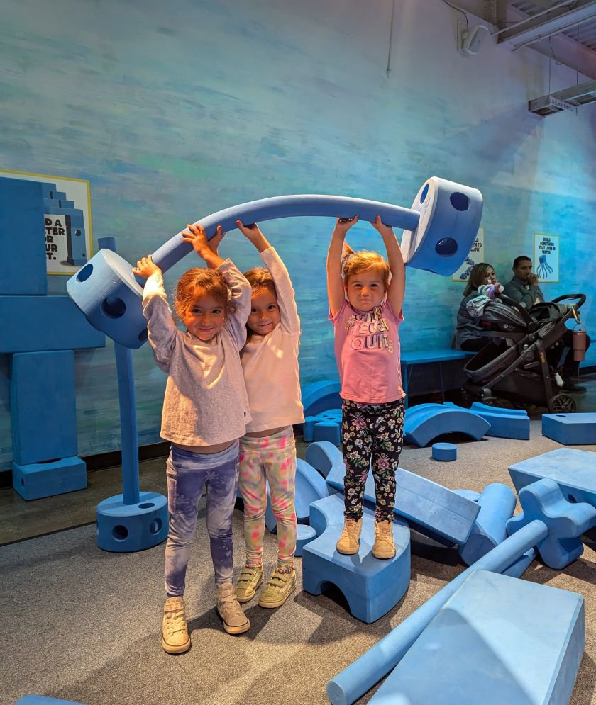 Three girls holding large blue foam 'blocks' mimicing a dumbbell above their heads. 