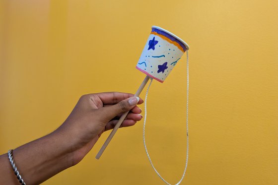 A person holding a dowel with a paper cup attached to it and a string with a ball at the end of the string. 