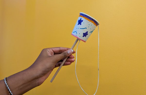 A person holding a dowel with a paper cup attached to it and a string with a ball at the end of the string. 