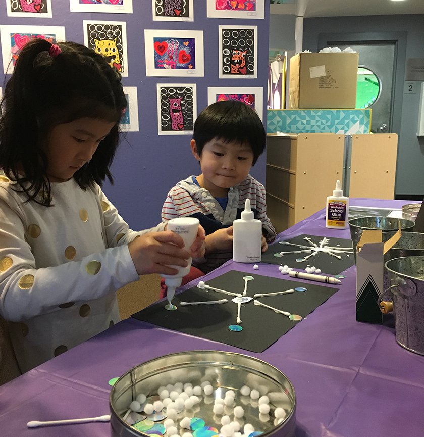 Two children holding glue making snowflakes out of Q-tips, and white pompoms. 