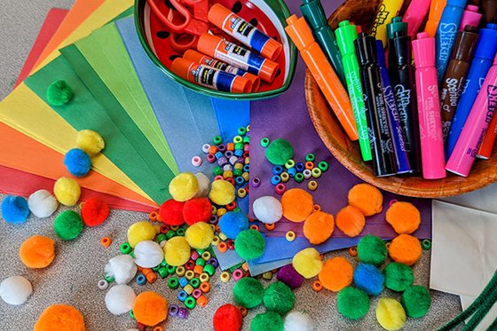A variety of colored tissue paper, beads, pompoms, markers, green pipe cleaners, and glue sticks spread across a table. 