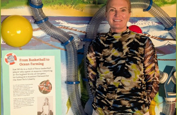 Sue Wicks stands inside museum exhibit, next to poster titled "From Basketball to Ocean Farming".