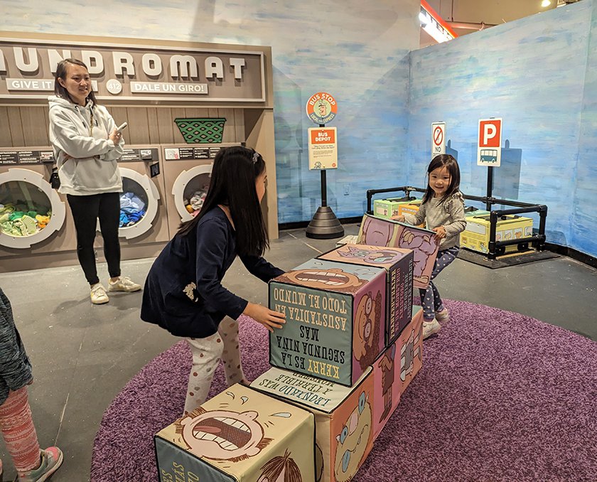 Two children with their adult in the Mo Willems exhibit stacking large soft blocks with images and phrases from Mo Willems books on them. 