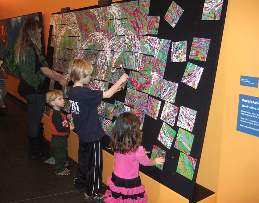 Children and an adult moving square pieces of painted canvas in order to connect white lines throughout painting. 