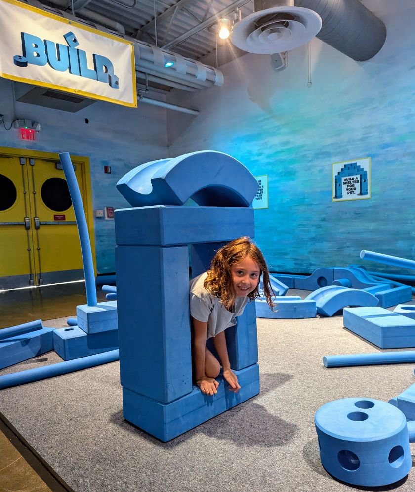 A child kneeling inside a structure made from large blue foam blocks while looking at the camera and smiling. 