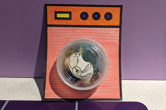 A paper washing machine with small a plastic container as the tub filled with pieces of fabric and a toy bunny. 