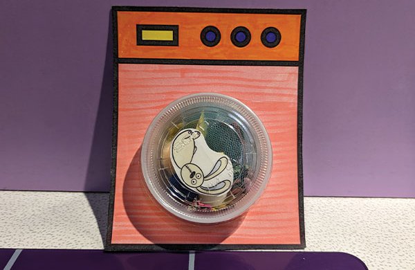 A paper washing machine with small a plastic container as the tub filled with pieces of fabric and a toy bunny. 