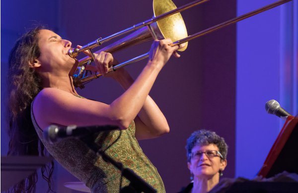 Woman in green sleeveless dress plays trombone. Grey haired woman watching sitting behind her. 