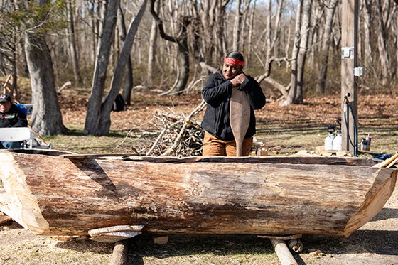 A traditional mishoon boat with a person standing in the background holding an oar. 