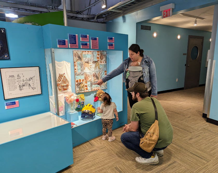 A family with two adults, a baby and a toddler looking at art behind plexiglass. 