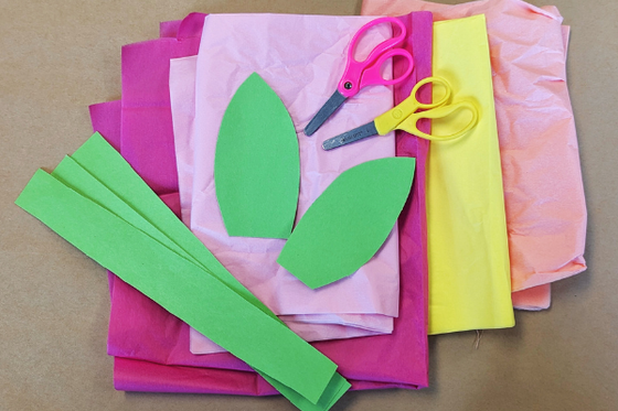 A stack of staggered pink and yellow tissue paper, scissors, and green pieces of construction paper. 