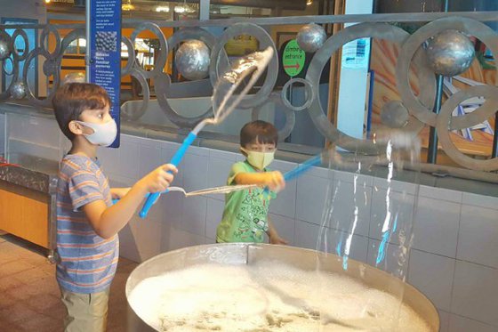 A child wearing a mask in the Bubbles exhibit holding a bubble wand making a bubble. 