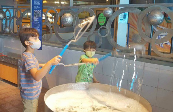 A child wearing a mask in the Bubbles exhibit holding a bubble wand making a bubble. 