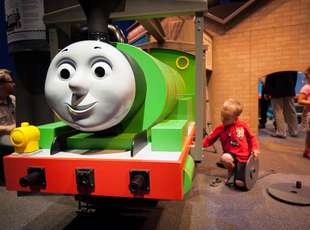 A child kneeling next to a large replica of Percy the green train changing the wheels. 