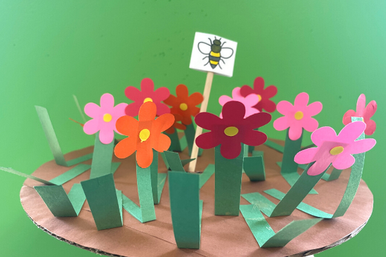 A round piece of cardboard decorated with paper flowers, blades of grass and a bee. 