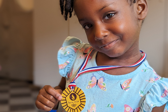 A child smiling at the camera while holding a paper gold medal around her neck. 