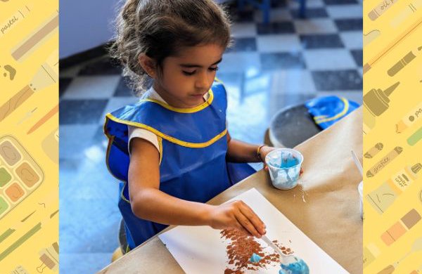 A child sitting at a table painting while wearing a blue smock with graphic images of paint, paintbrushes, pastels and colored pencils in the background. 