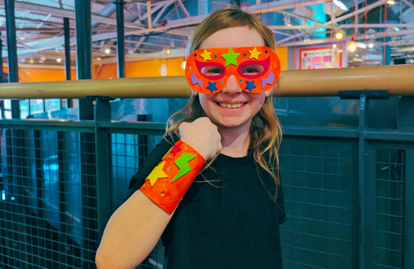 A child looking into the camera smiling while wearing an orange superhero mask and wrist guard decorated with multicolored stars and lightening bolts.