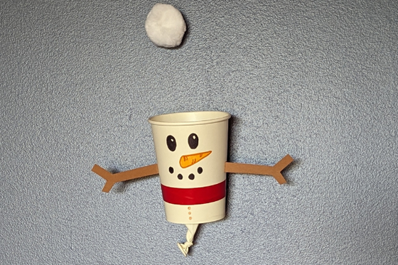A white cup decroated as a snowperson with brown arms, a white balloon tied to the bottom, and a cotton ball above. 