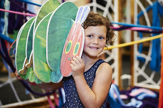 A child smiling while holding a wooden Very Hungry Caterpillar toy against their face. 
