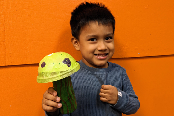 A child against a orange wall smiling and holding a colorful mushroom made with a toilet paper roll and rounded cup cover. 