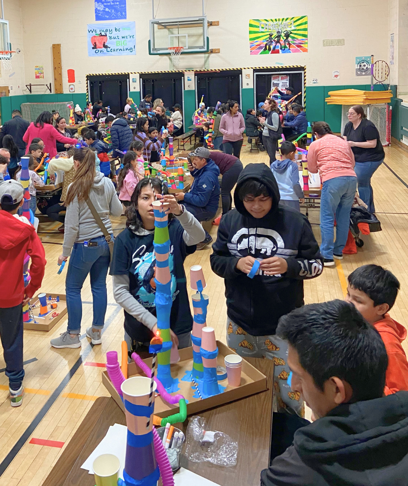 Children and adults standing at tables building rollercoasters from cups, straws and tubes. 