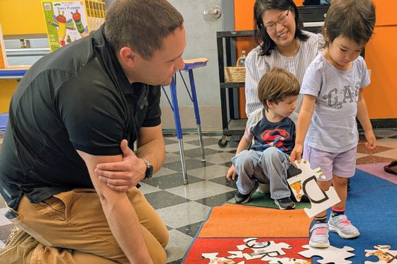 Child engages with puzzle on carpet with educator next and caregiver behind. 