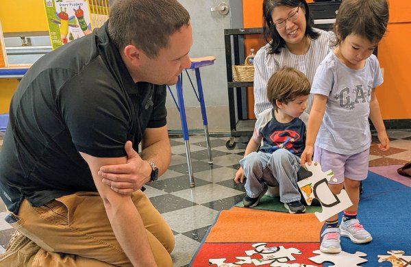 Child engages with puzzle on carpet with educator next and caregiver behind. 