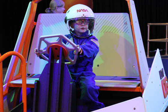 A child wearing a space suit and a white helmet with NASA logo driving a rover. 