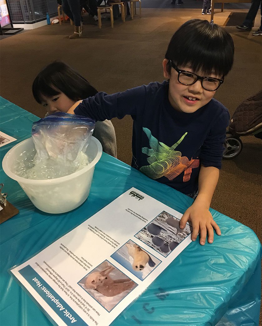 Two children standing in front of a table and sticking their hand in a bowl of ice water. 
